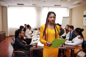 Face of handsome african business woman in yellow dress, holding clipboard on the background of business peoples multiracial team meeting, sitting in office table.