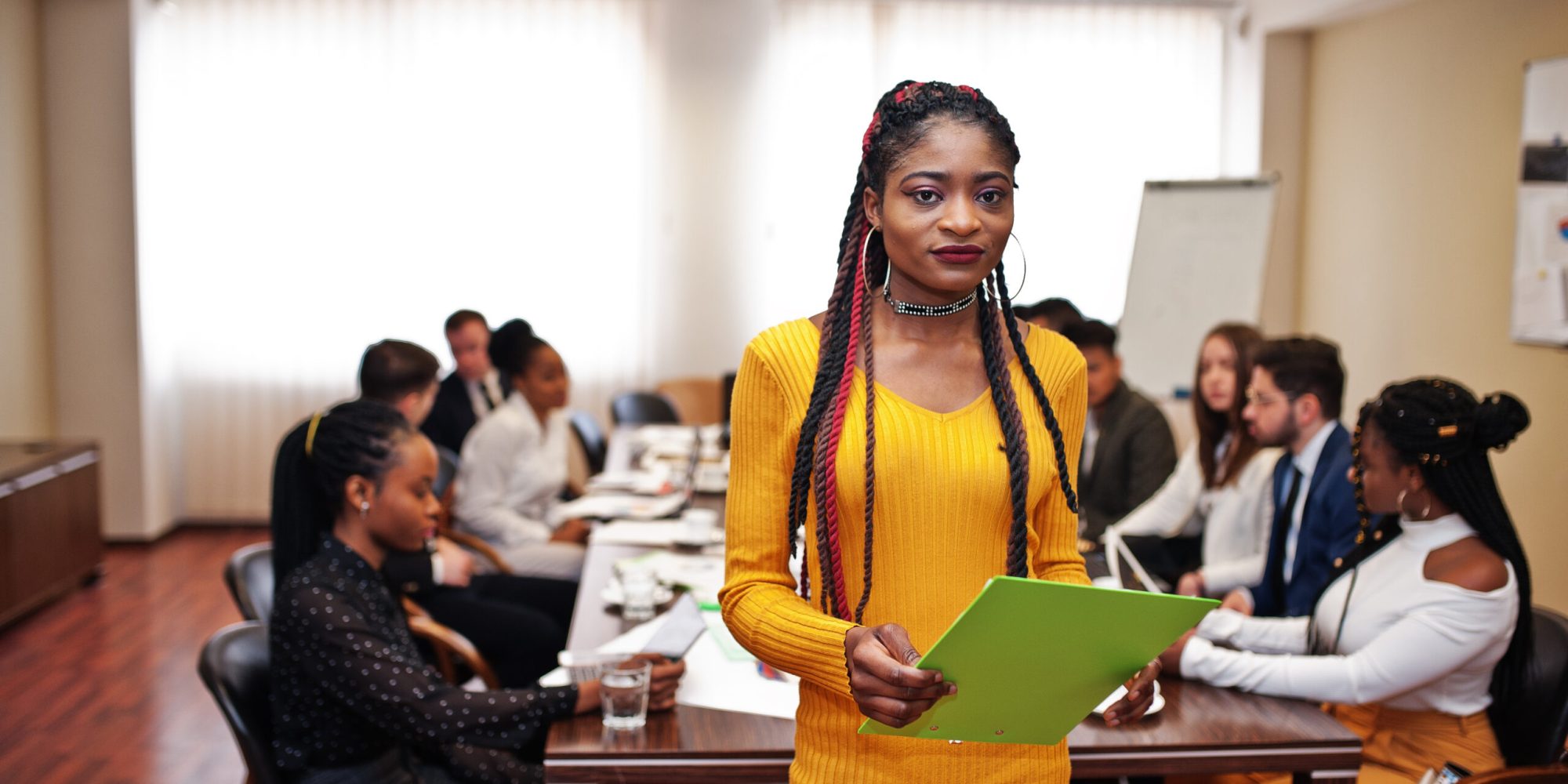 Face of handsome african business woman in yellow dress, holding clipboard on the background of business peoples multiracial team meeting, sitting in office table.